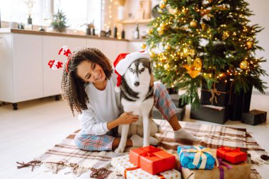 Mutlu bir kadın etrafta hediyelerle Noel ağacının yanında otururken köpeğiyle oynayıp eğleniyor. Kış tatili Yeni Yıl.