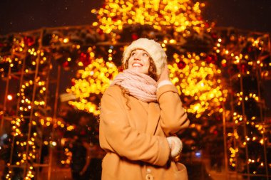 Kış tarzı kıyafetler giymiş genç bir kadın akşamları çelenk ışıklarına karşı. Soğuk hava. Kış modası, tatiller, dinlenme, seyahat konsepti.