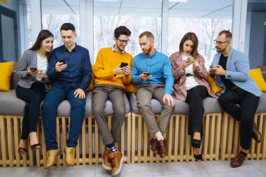 Cep telefonu kullanan bir grup genç. Modern bir ofiste dijital cihazlarla yan yana oturan iş kolejleri. Öğrenim, iş, teknoloji.