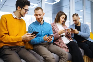 Cep telefonu kullanan bir grup genç. Modern bir ofiste dijital cihazlarla yan yana oturan iş kolejleri. Öğrenim, iş, teknoloji.