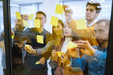 Genç iş adamları cam duvar işaretleri ve çıkartmaların önünde strateji tartışıp planlıyorlar. Başlangıç ofisi. Beyin fırtınası konsepti. Eğitim, analiz.