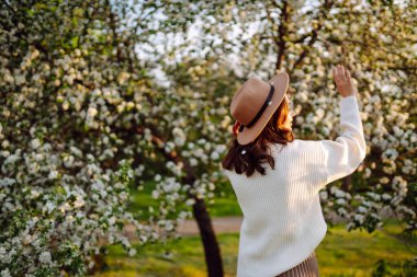 Çiçek açan bahar bahçesinde kokunun tadını çıkaran genç bir kadın. Rahatlama, seyahat, özgürlük ve bahar tatili kavramı. Moda tarzı