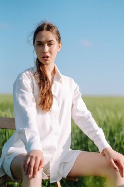 Beyaz takım elbiseli genç kadın yeşil buğday tarlasında poz veriyor. Moda ve yaşam tarzı konsepti. İnsanlar, seyahat, doğa ve tatil kavramı.