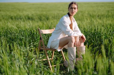 Beyaz takım elbiseli genç kadın yeşil buğday tarlasında poz veriyor. Moda ve yaşam tarzı konsepti. İnsanlar, seyahat, doğa ve tatil kavramı.