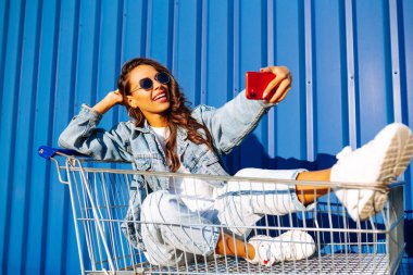 Selfie time. Young woman  having fun on shopping trolleys. Consumerism, sale, discounts, lifestyle concept.
