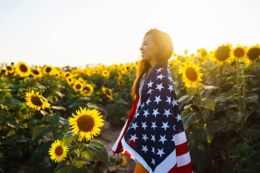 Ayçiçeği tarlasında Amerikan bayrağını sallayan genç bir kadın. Vatansever, gün batımında ulusal Amerikan bayrağını göndere çekiyor. Bağımsızlık Günü, 4 Temmuz.