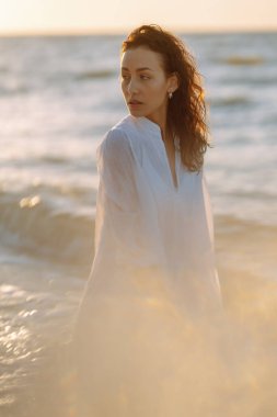 Beyaz kanat çırpan elbiseli genç ve mutlu bir kadın deniz kıyısında yürüyor. Kız sihirli gün doğumuna bakıyor. Seyahat, hafta sonu, rahatlama ve yaşam tarzı konsepti.