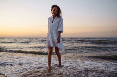 Beyaz kanat çırpan elbiseli genç ve mutlu bir kadın deniz kıyısında yürüyor. Kız sihirli gün doğumuna bakıyor. Seyahat, hafta sonu, rahatlama ve yaşam tarzı konsepti.