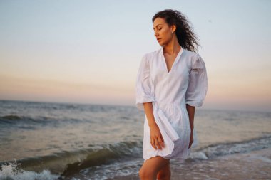 Beyaz kanat çırpan elbiseli genç ve mutlu bir kadın deniz kıyısında yürüyor. Kız sihirli gün doğumuna bakıyor. Seyahat, hafta sonu, rahatlama ve yaşam tarzı konsepti.