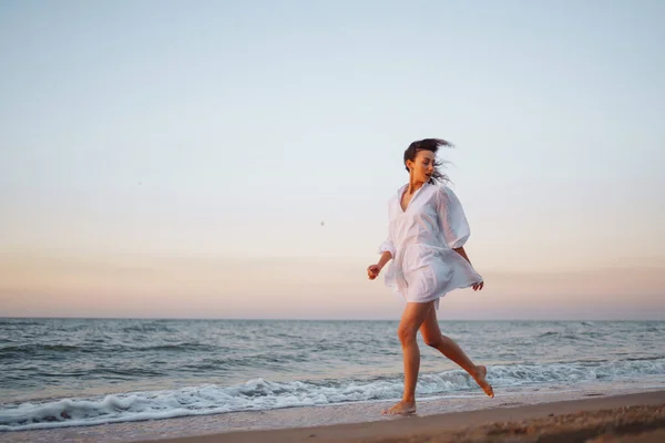白いなびくドレスを着た若い幸せな女性が海岸沿いを歩いている 少女は不思議な日の出を見る リラックスしてライフスタイルのコンセプト — ストック写真