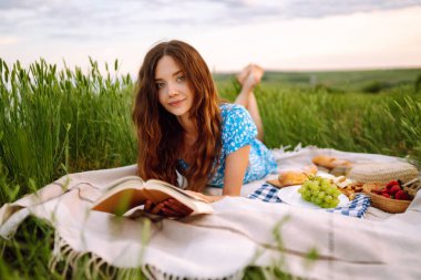 Genç bir kadın elinde bir kitapla ekose üzerinde oturuyor. Doğada yaz pikniği. Sağlıklı yiyecekler. İnsanlar, yaşam tarzı, rahatlama ve tatil kavramı.