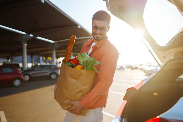 Shopping time. Healthy vegan vegetarian food in a paper bag in the male hands. Young man with shopping bag full of vegetables near the car.