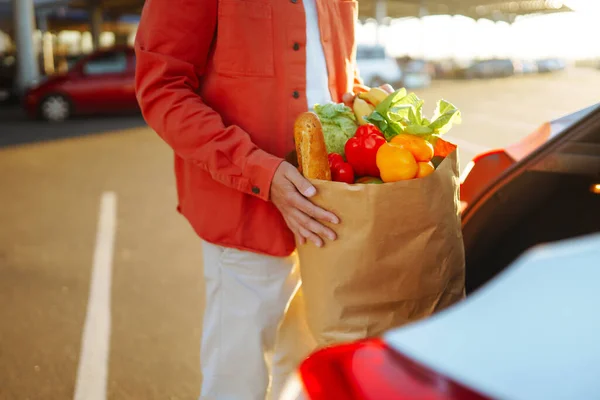 Shopping time. Healthy vegan vegetarian food in a paper bag in the male hands. Young man with shopping bag full of vegetables near the car.