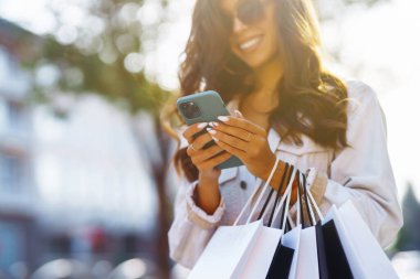 Alışveriş çantalı genç bir kadın şehir caddesinde telefonunu kullanıyor. Satın almalar, kara cuma, indirimler, satış konsepti. Çevrimiçi alışveriş kavramı.