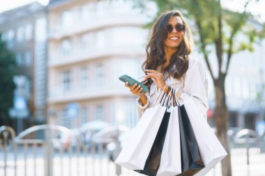 Alışveriş çantalı genç bir kadın şehir caddesinde telefonunu kullanıyor. Satın almalar, kara cuma, indirimler, satış konsepti. Çevrimiçi alışveriş kavramı.