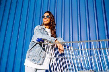 Alışveriş arabalı mutlu kadın renk duvarının yanında eğleniyor. Tüketim, satış, alım, alışveriş, yaşam tarzı konsepti.