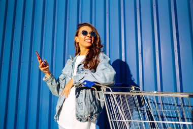 Alışveriş arabalı mutlu kadın renk duvarının yanında eğleniyor. Tüketim, satış, alım, alışveriş, yaşam tarzı konsepti.