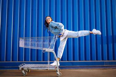 Alışveriş arabalı mutlu kadın renk duvarının yanında eğleniyor. Tüketim, satış, alım, alışveriş, yaşam tarzı konsepti.