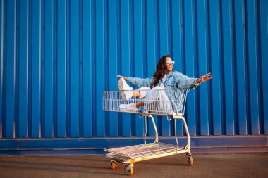 Alışveriş arabalı mutlu kadın renk duvarının yanında eğleniyor. Tüketim, satış, alım, alışveriş, yaşam tarzı konsepti.