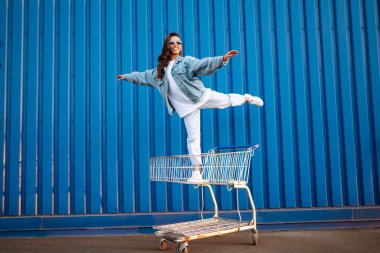 Alışveriş arabalı mutlu kadın renk duvarının yanında eğleniyor. Tüketim, satış, alım, alışveriş, yaşam tarzı konsepti.