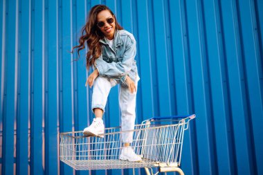 Alışveriş arabalı mutlu kadın renk duvarının yanında eğleniyor. Tüketim, satış, alım, alışveriş, yaşam tarzı konsepti.