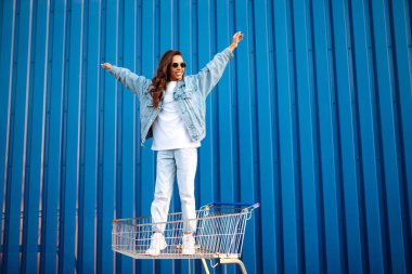 Alışveriş arabalı mutlu kadın renk duvarının yanında eğleniyor. Tüketim, satış, alım, alışveriş, yaşam tarzı konsepti.