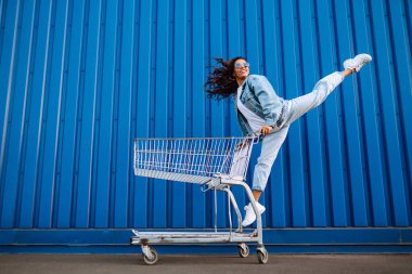 Alışveriş arabalı mutlu kadın renk duvarının yanında eğleniyor. Tüketim, satış, alım, alışveriş, yaşam tarzı konsepti.