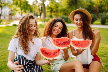 Üç genç kadın birlikte eğleniyor ve sıcak yaz gününde karpuz yiyor. İnsanlar, yaşam tarzı, seyahat, doğa ve tatil kavramı. 