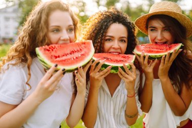 Üç genç kadın birlikte eğleniyor ve sıcak yaz gününde karpuz yiyor. İnsanlar, yaşam tarzı, seyahat, doğa ve tatil kavramı. 