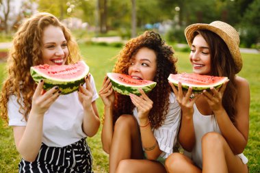 Üç genç kadın birlikte eğleniyor ve sıcak yaz gününde karpuz yiyor. İnsanlar, yaşam tarzı, seyahat, doğa ve tatil kavramı. 