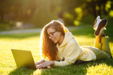 İnternette kablosuz kulaklıklı genç bir kadın çalışıyor ya da parkta çimlerde oturuyor. İş, blog yazma, serbest çalışma, eğitim kavramı.