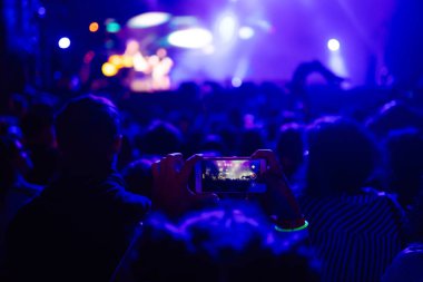 Using a smartphone in a public event, live music festival. Holding a mobile phone in hands and shooting photo or video content. Youth, party, vacation concept.