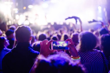 Using a smartphone in a public event, live music festival. Holding a mobile phone in hands and shooting photo or video content. Youth, party, vacation concept.