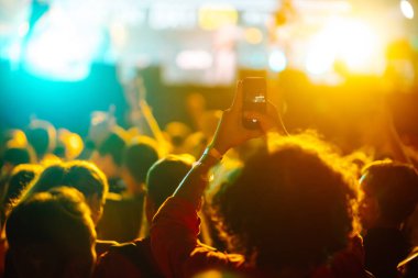 Using a smartphone in a public event, live music festival. Holding a mobile phone in hands and shooting photo or video content. Youth, party, vacation concept.