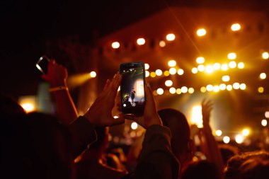 Using a smartphone in a public event, live music festival. Holding a mobile phone in hands and shooting photo or video content. Youth, party, vacation concept.