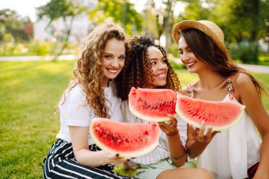 Three young woman  camping on the grass, eating watermelon, laughing. People, lifestyle, travel, nature and vacations concept.