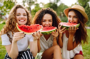 Three young woman  camping on the grass, eating watermelon, laughing. People, lifestyle, travel, nature and vacations concept.