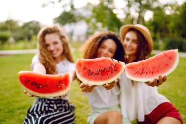 Three young woman  camping on the grass, eating watermelon, laughing. People, lifestyle, travel, nature and vacations concept.