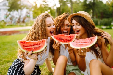 Three young woman  camping on the grass, eating watermelon, laughing. People, lifestyle, travel, nature and vacations concept.
