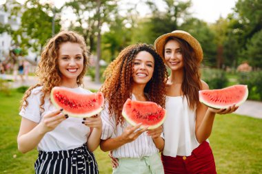 Three young woman  camping on the grass, eating watermelon, laughing. People, lifestyle, travel, nature and vacations concept.