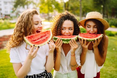 Three young woman  camping on the grass, eating watermelon, laughing. People, lifestyle, travel, nature and vacations concept.