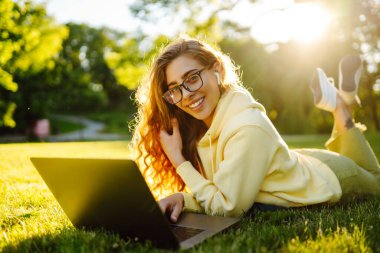 Beautiful woman  working online or studying at laptop sitting on grass at park. Business, blogging, freelance, education concept.