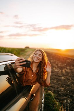 Selfie zamanı. Genç bir kadın arabada selfie çekiyor ve günbatımının tadını çıkarıyor. Yaşam tarzı, seyahat, turizm, doğa, aktif yaşam.