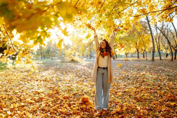 Young woman in park on sunny autumn day, smiling, having fun with leaves. Autumn fashion. Lifestyle. Relax, nature concept.