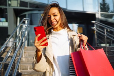 Yolda elinde poşetlerle dikilen ve cep telefonu kullanan genç bir kadın. İnternetten alışveriş konsepti. Satın almalar, Kara Cuma, indirimler, satış konsepti.