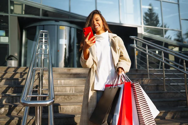 若い女性は 道路に沿ってショッピングバッグを保持し 携帯電話を使用して立っている オンラインショッピングのコンセプト 黒の金曜日 販売の概念 — ストック写真