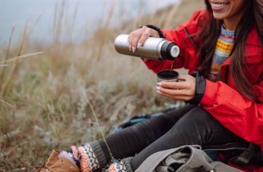 Genç kadın bir termos tutuyor ve sonbahar ormanında çay içiyor. Dinlenme, rahatlama, seyahat, yaşam tarzı konsepti.