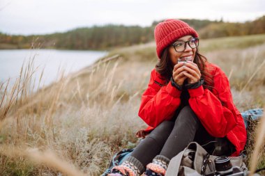 Genç kadın bir termos tutuyor ve sonbahar ormanında çay içiyor. Dinlenme, rahatlama, seyahat, yaşam tarzı konsepti.