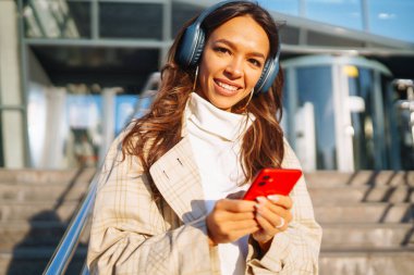 Genç ve güzel bir kadın kulaklıkla müzik dinlerken şehirde cep telefonu kullanıyor. İnsanların, müziğin, duyguların konsepti.