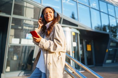 Genç ve güzel bir kadın kulaklıkla müzik dinlerken şehirde cep telefonu kullanıyor. İnsanların, müziğin, duyguların konsepti.